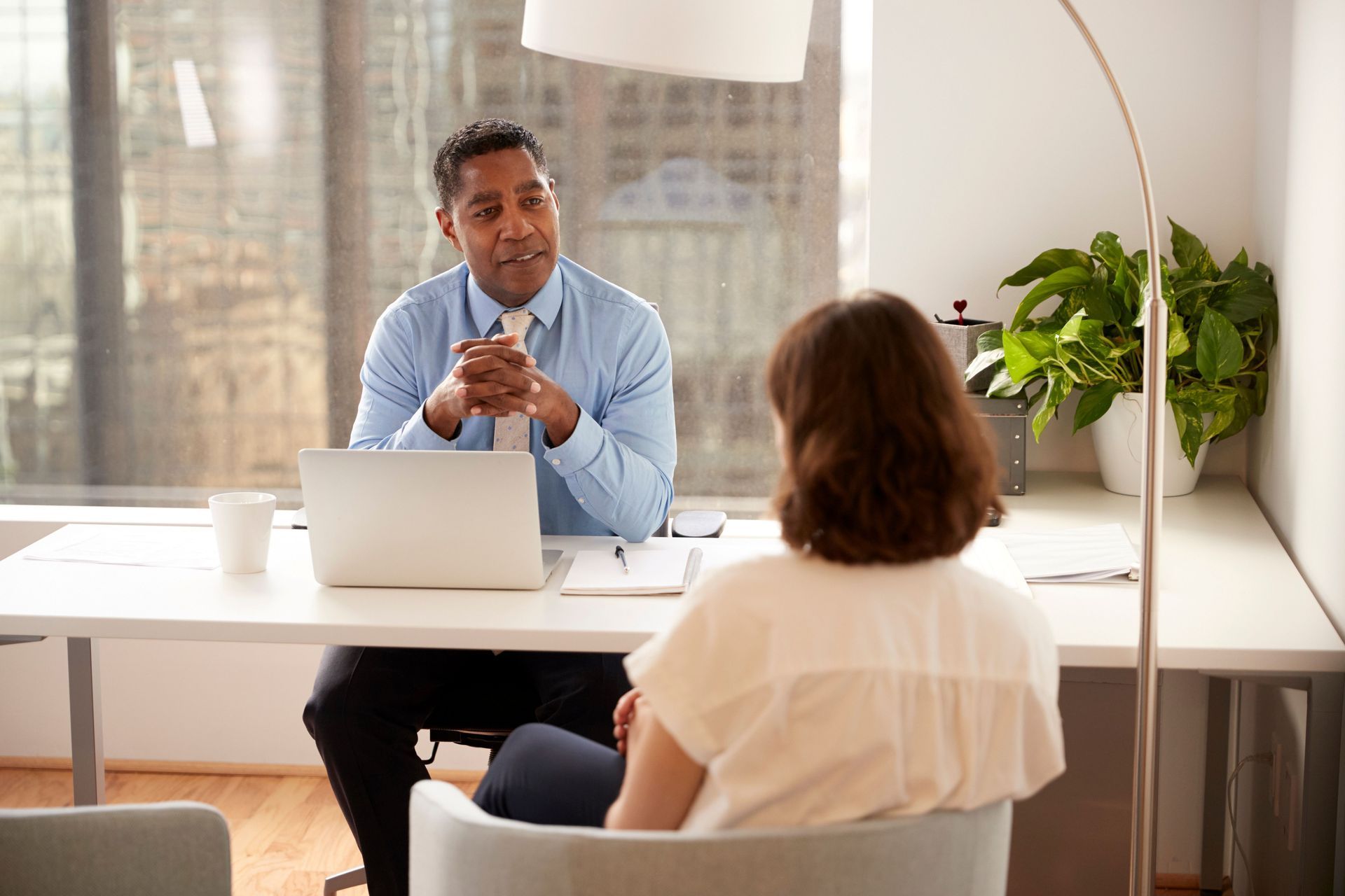 Man in blue shirt speaks to woman at desk with laptop in an office setting.