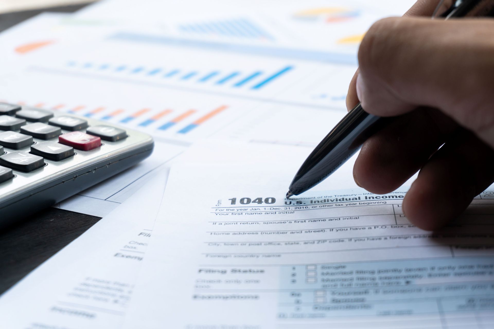 Hand writing on a 1040 tax form with a calculator and financial charts in the background.