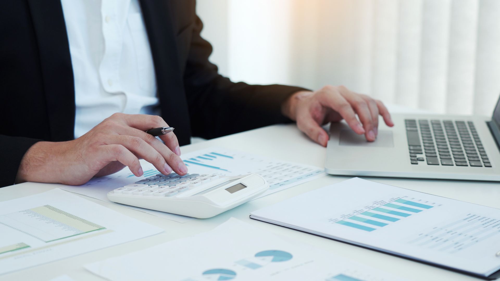 Person using a calculator and laptop, reviewing charts and graphs on a desk.
