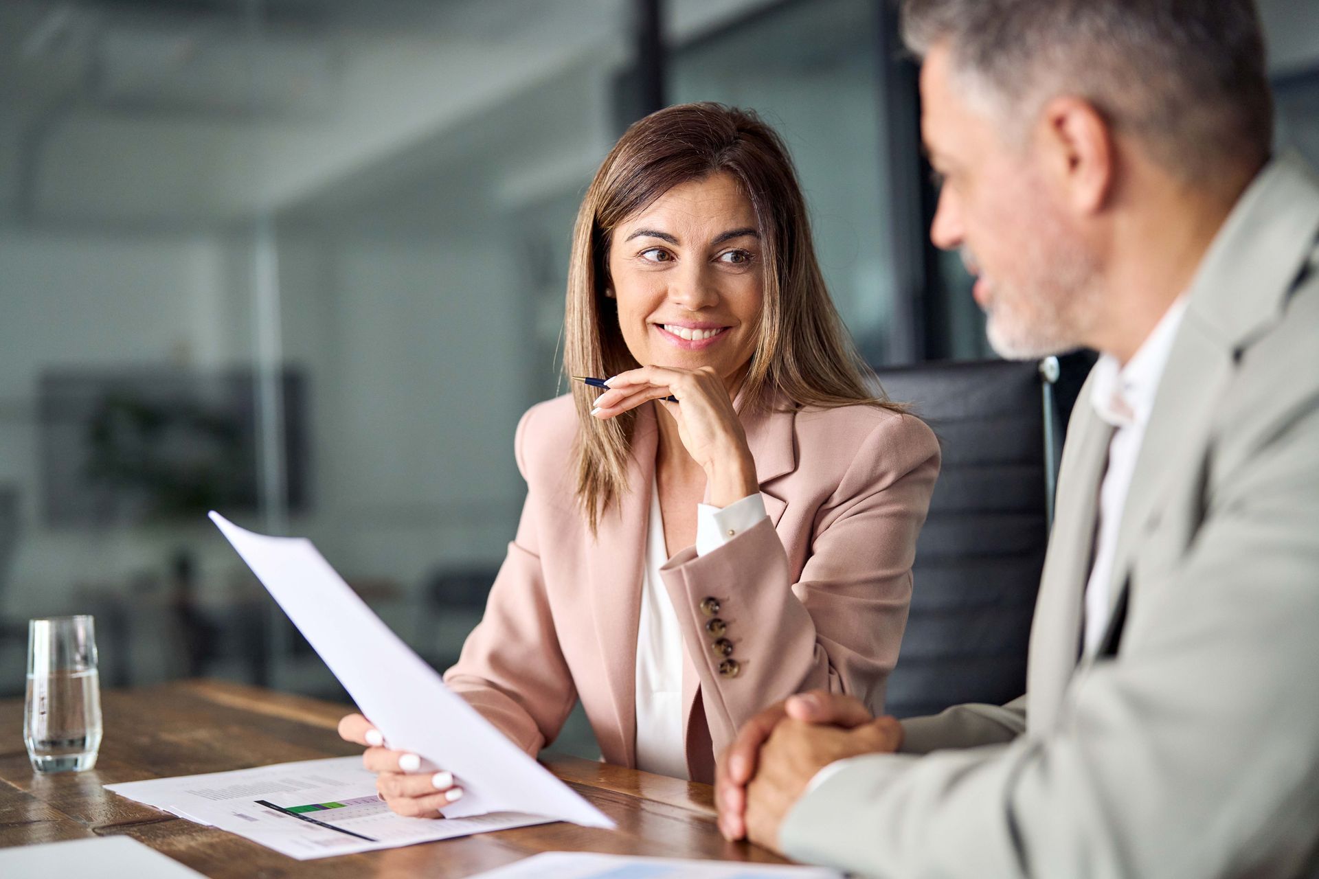 Woman in pink blazer smiles at a man, reviewing documents at a table.