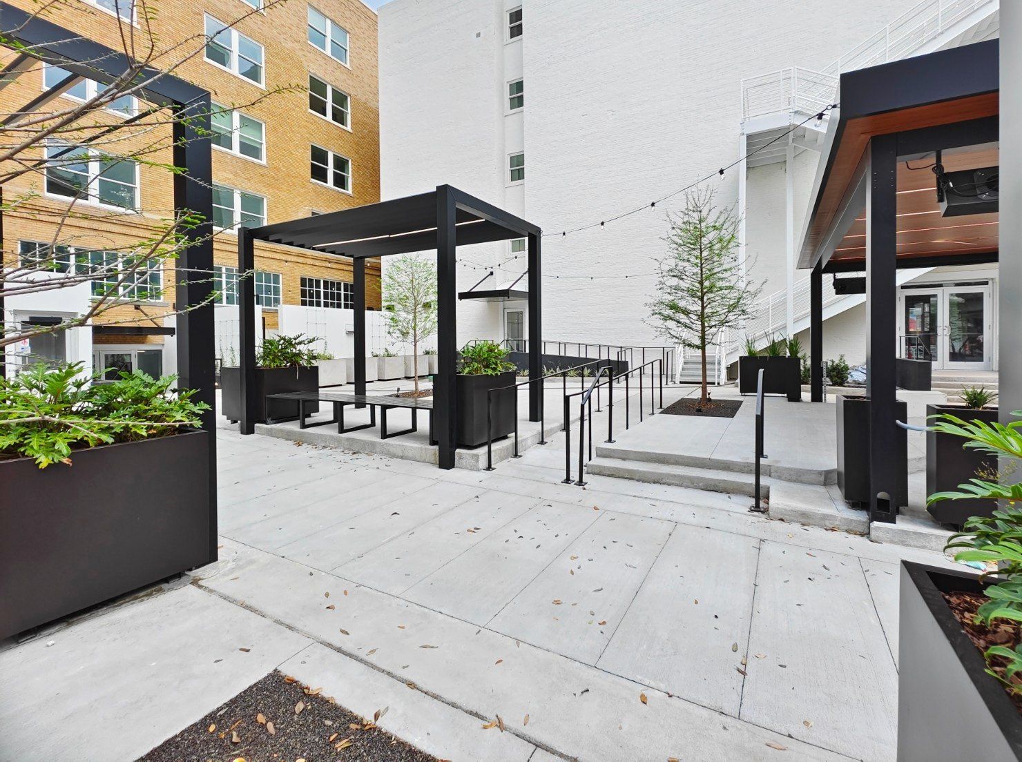 Outdoor courtyard with concrete walkways, black pergolas, planters, and trees.