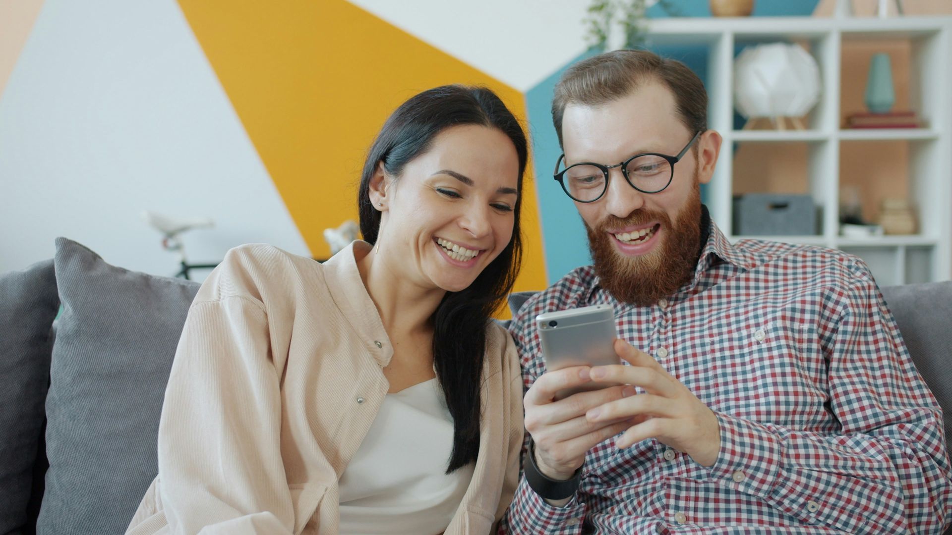 A smiling couple looks at a phone on a couch, indoors. The man has a beard and glasses.