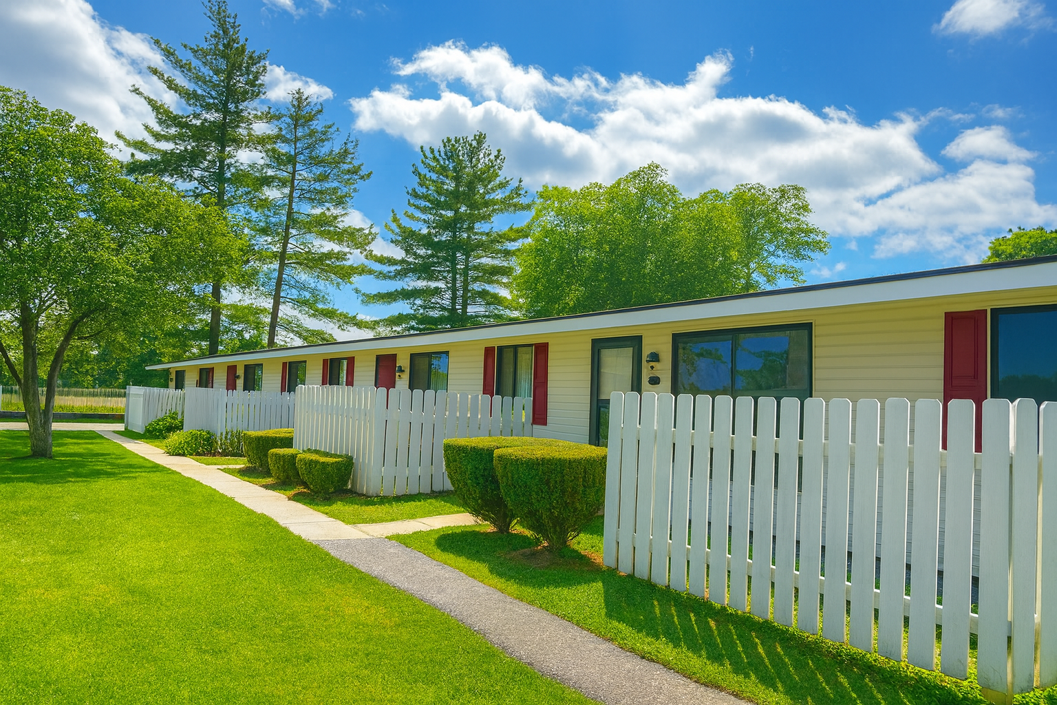 Row of light yellow townhouses with white picket fence, green grass, and blue sky.