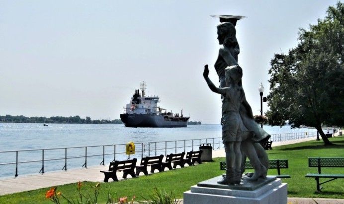 Statue of three figures overlooking water with a ship in the background. Green grass and benches in the foreground.