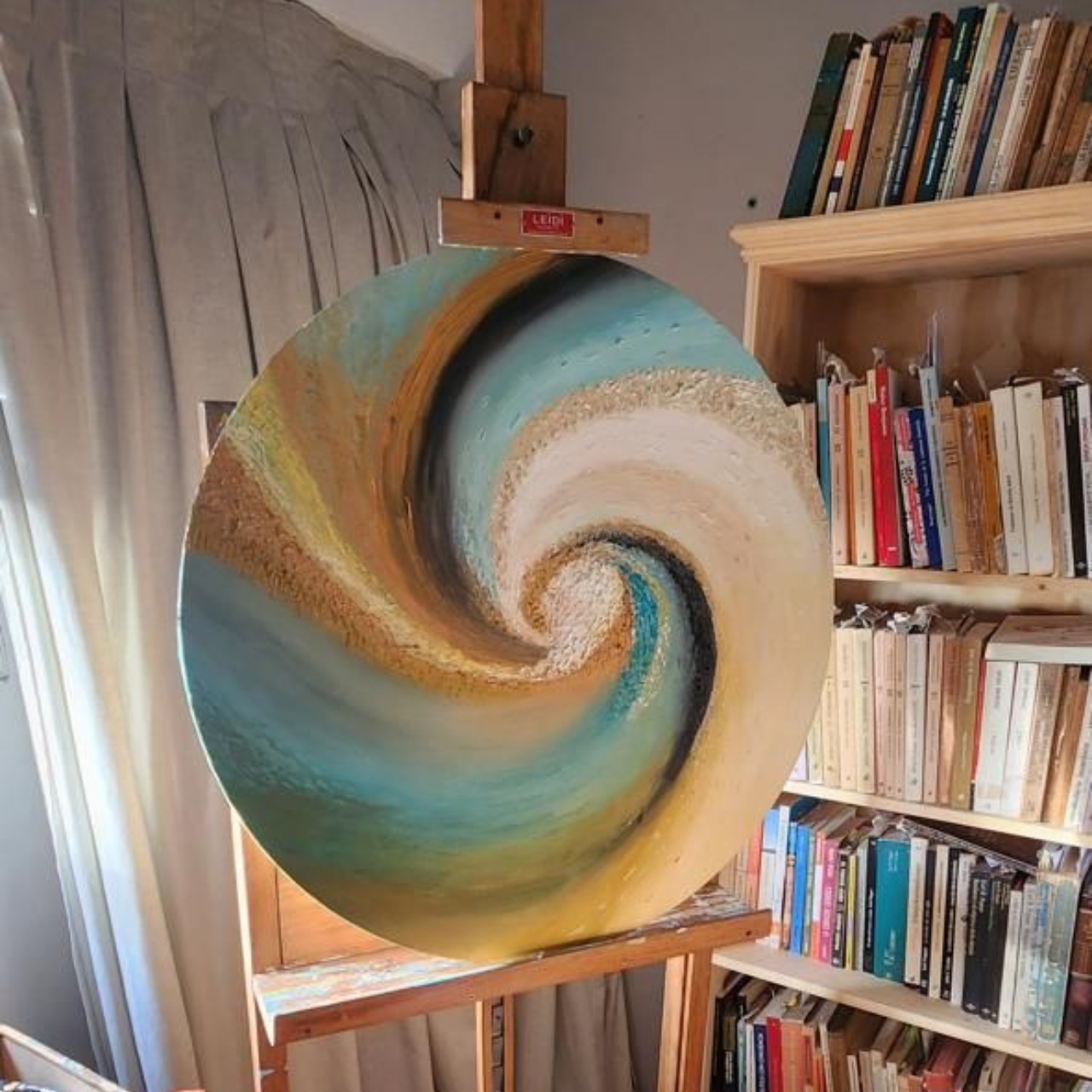 Pintura circular abstracta sobre caballete; diseño en espiral con dorado, azul, canela y blanco. Estantería al fondo.