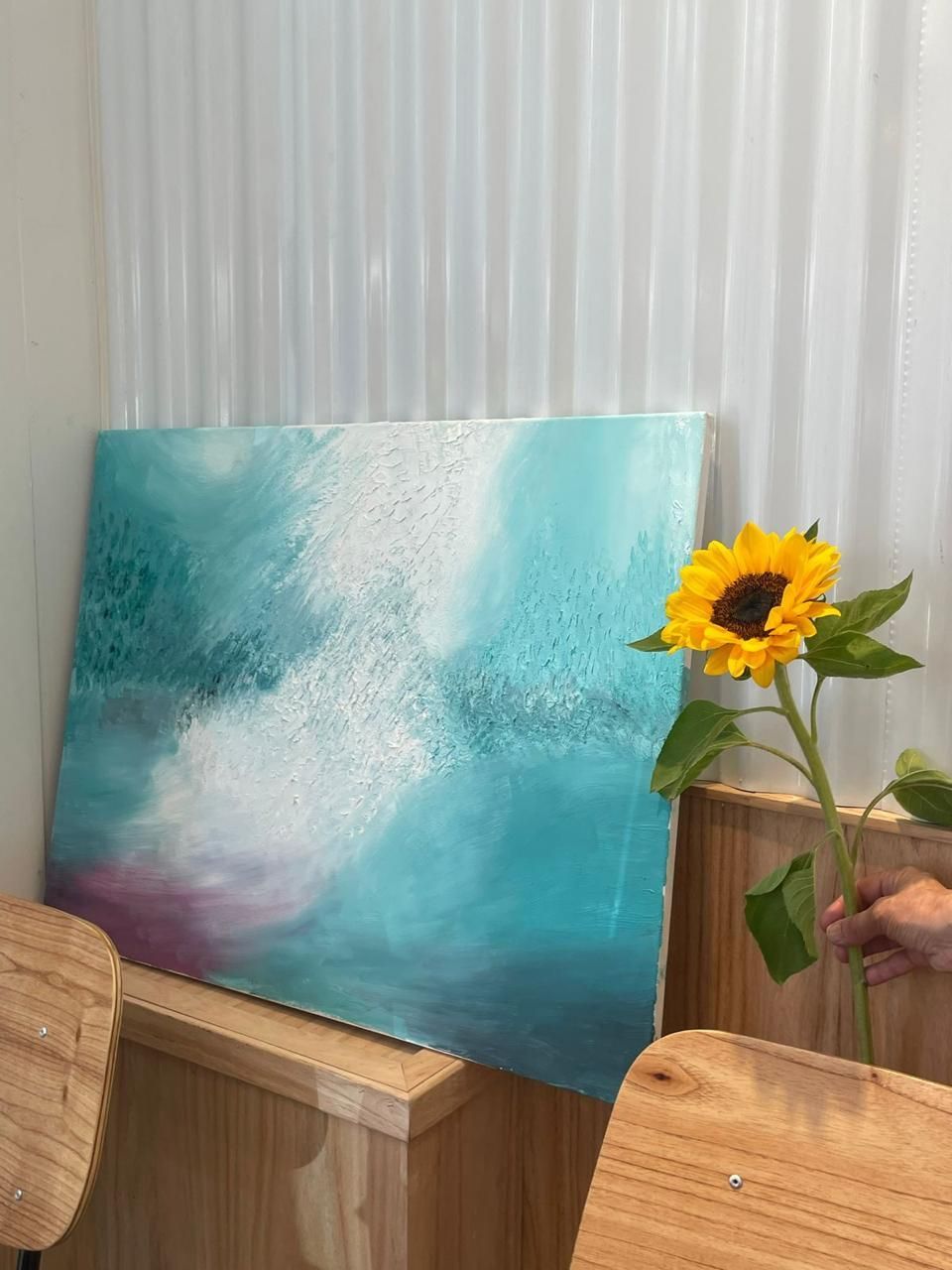 Cuadro abstracto en verde azulado, blanco y morado junto a un girasol sostenido por una persona.