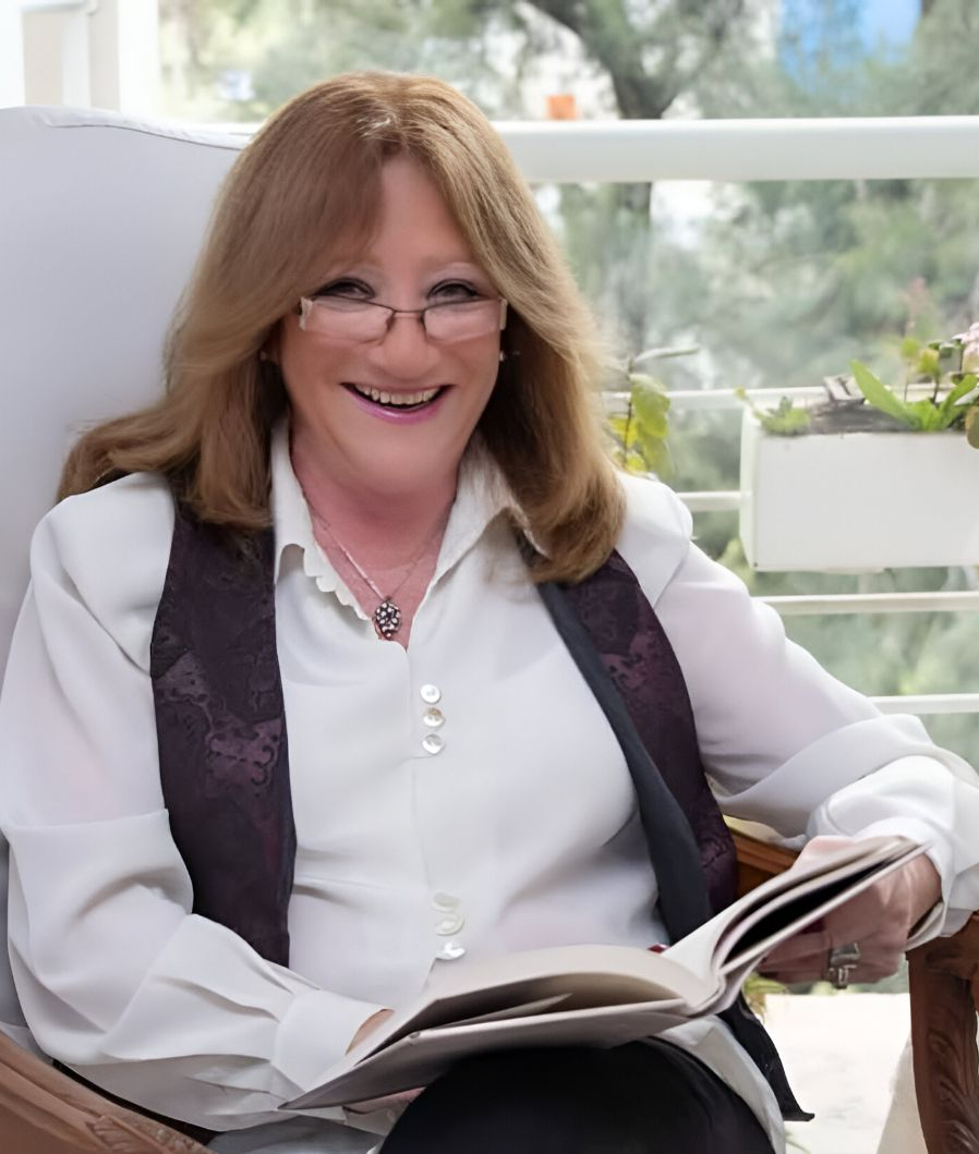 Mujer sentada leyendo un libro, con gafas y chaleco, sonriendo. Al aire libre.