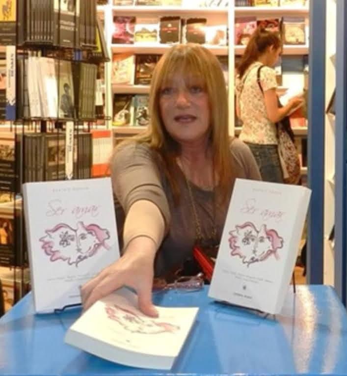 Una mujer sentada en una mesa de librería con libros. Coge uno con una ilustración rosa en la portada. Estantes al fondo.