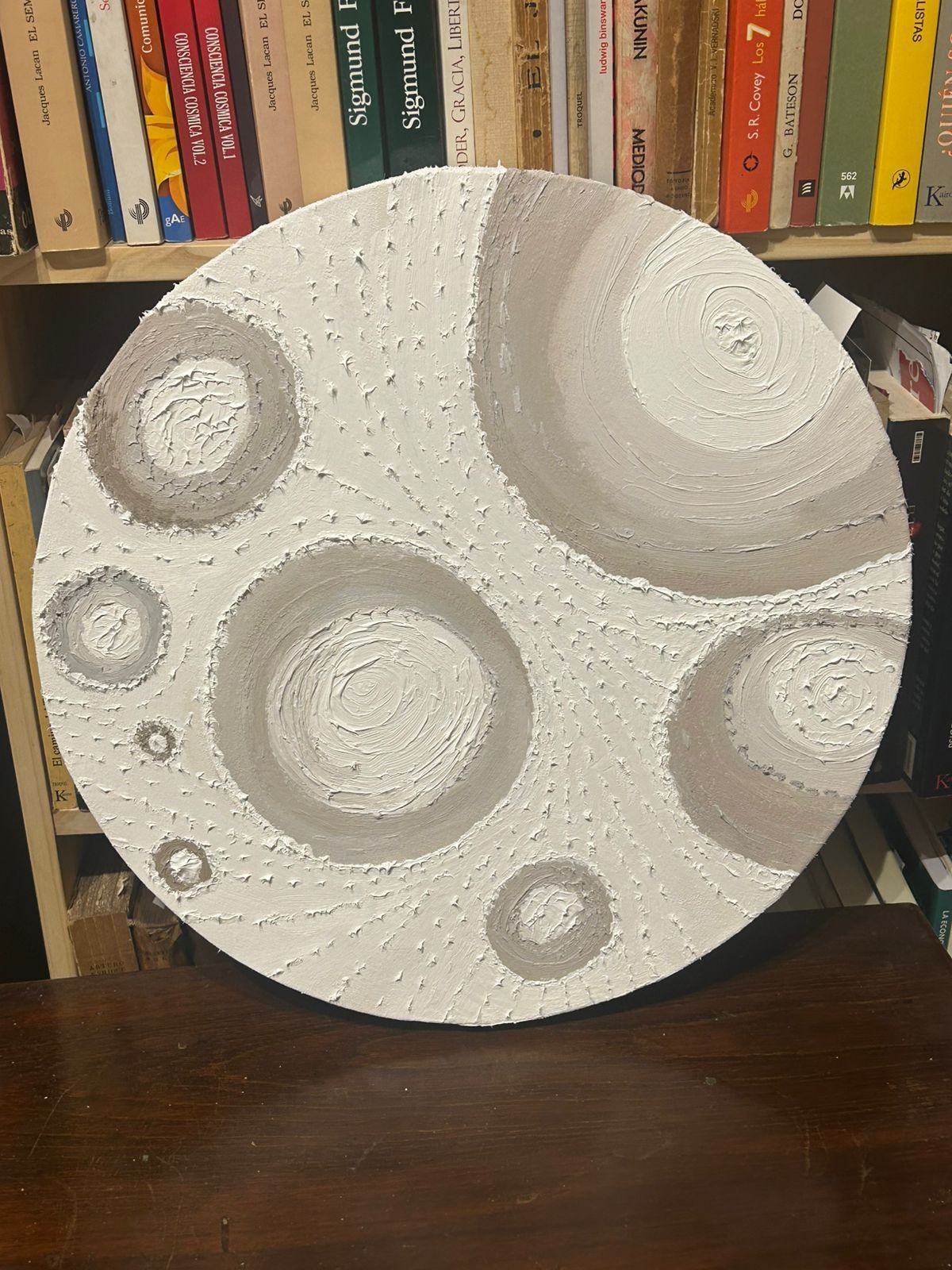 Cuadro circular con círculos de color gris claro sobre fondo blanco texturizado, colocado sobre una estantería.