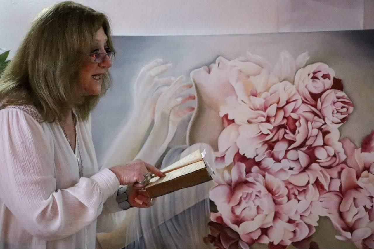 Mujer con gafas sosteniendo un libro, haciendo un gesto hacia una pintura de flores y una cara.
