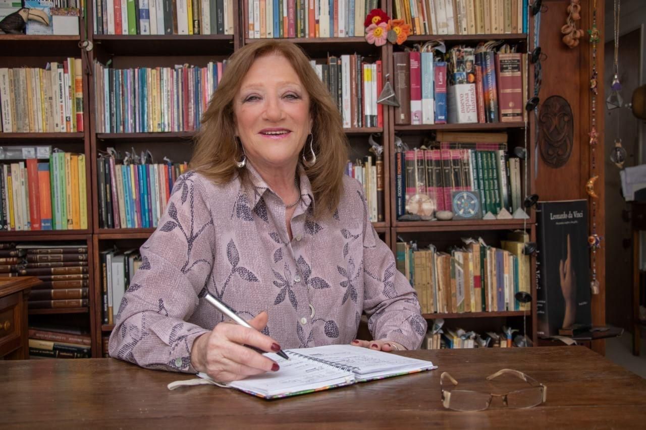 Mujer escribiendo en un cuaderno en el escritorio, estanterías en el fondo.