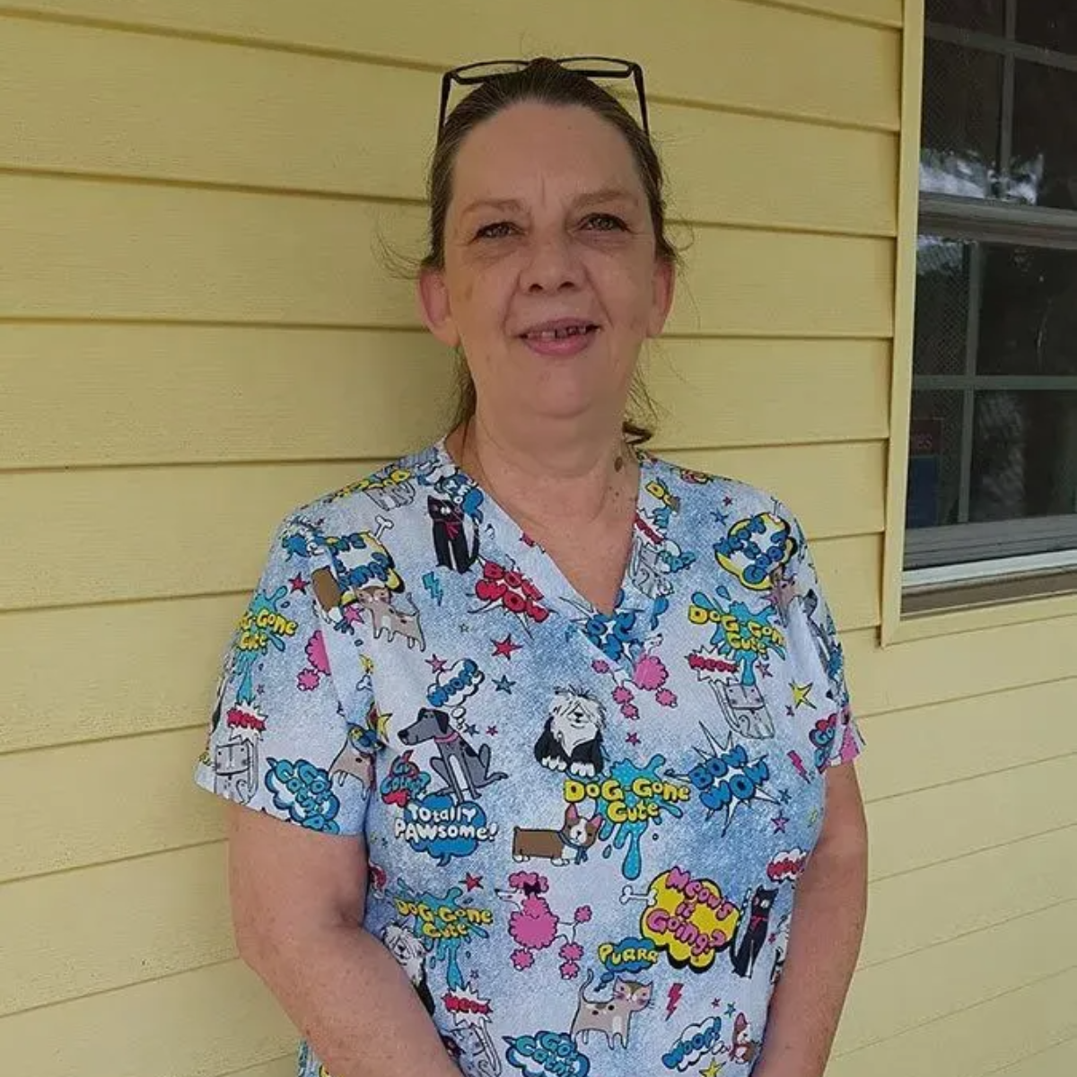 Woman in cartoon scrubs stands in front of a yellow wall with a window.