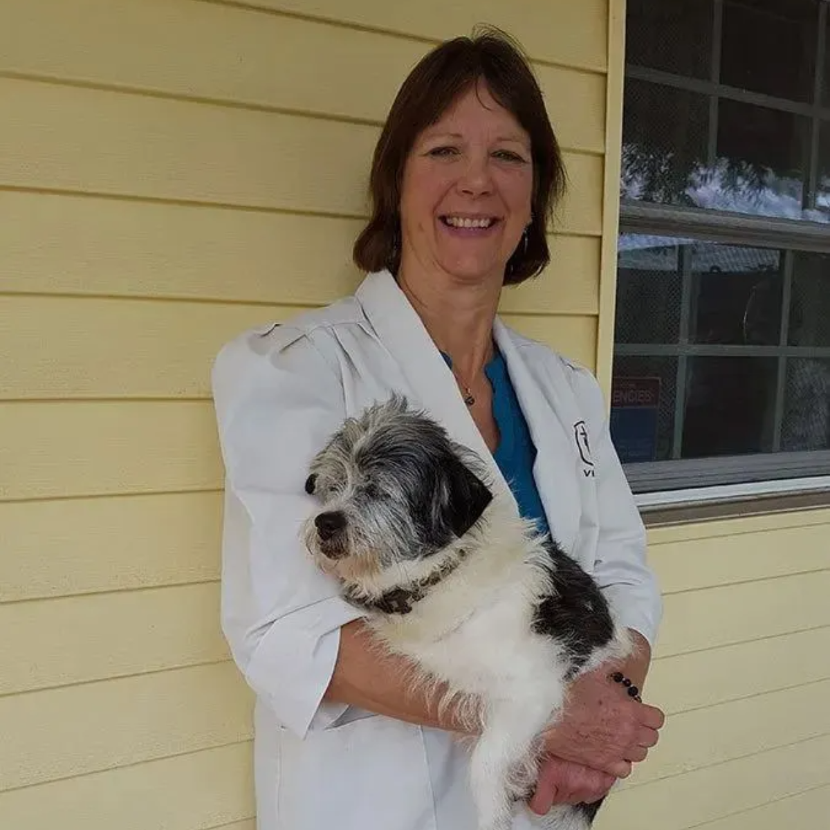 Woman in white coat holding small dog, smiling in front of a yellow building with a window.