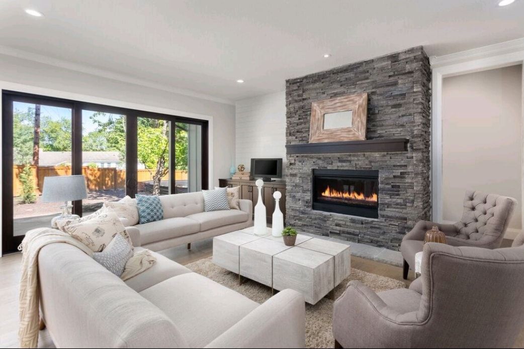 Bright modern living room with white sofas, stone fireplace, and large windows overlooking trees