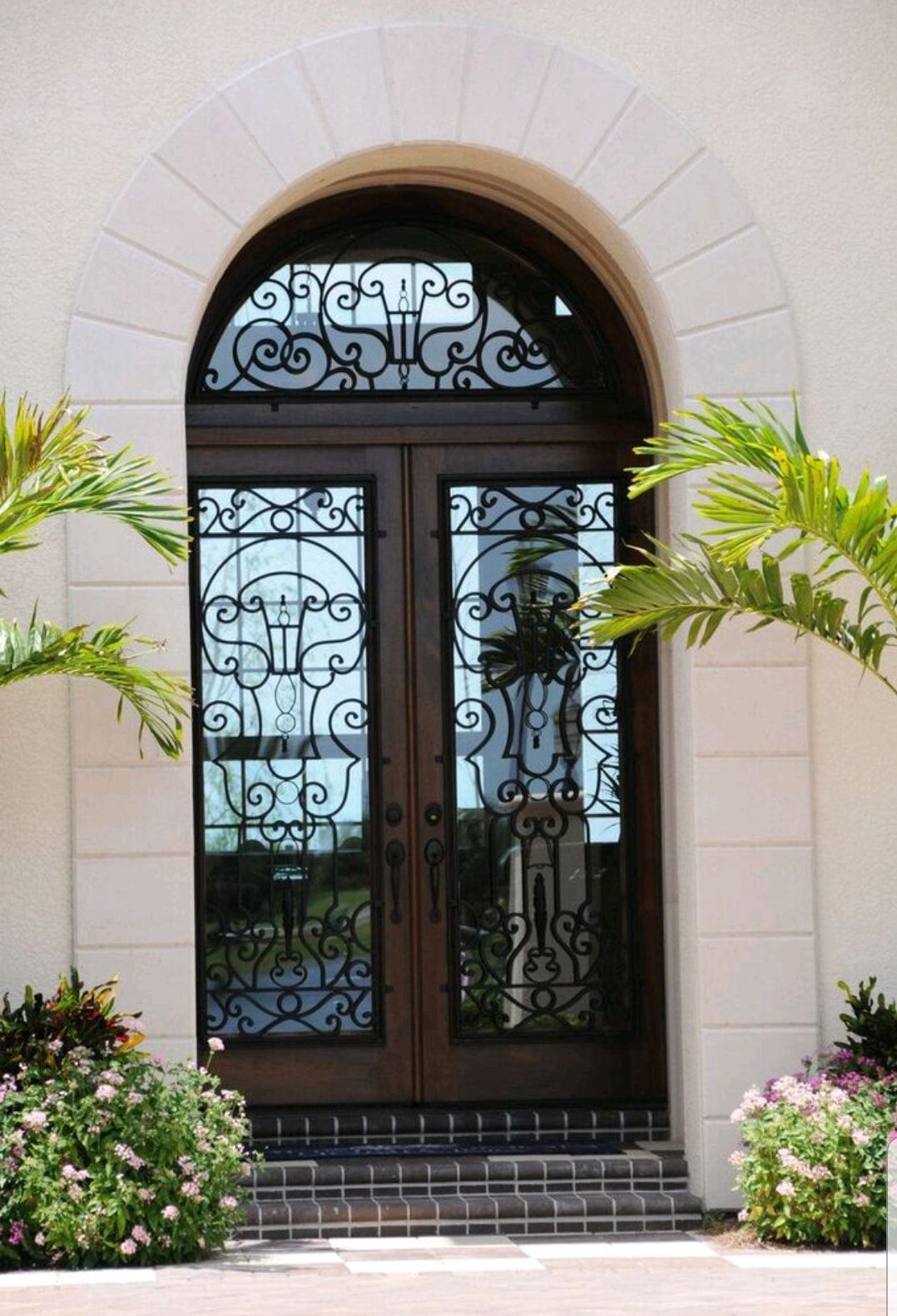 Arched double glass doors with ornate ironwork, framed by pale stone and flanked by palm plants and flowers