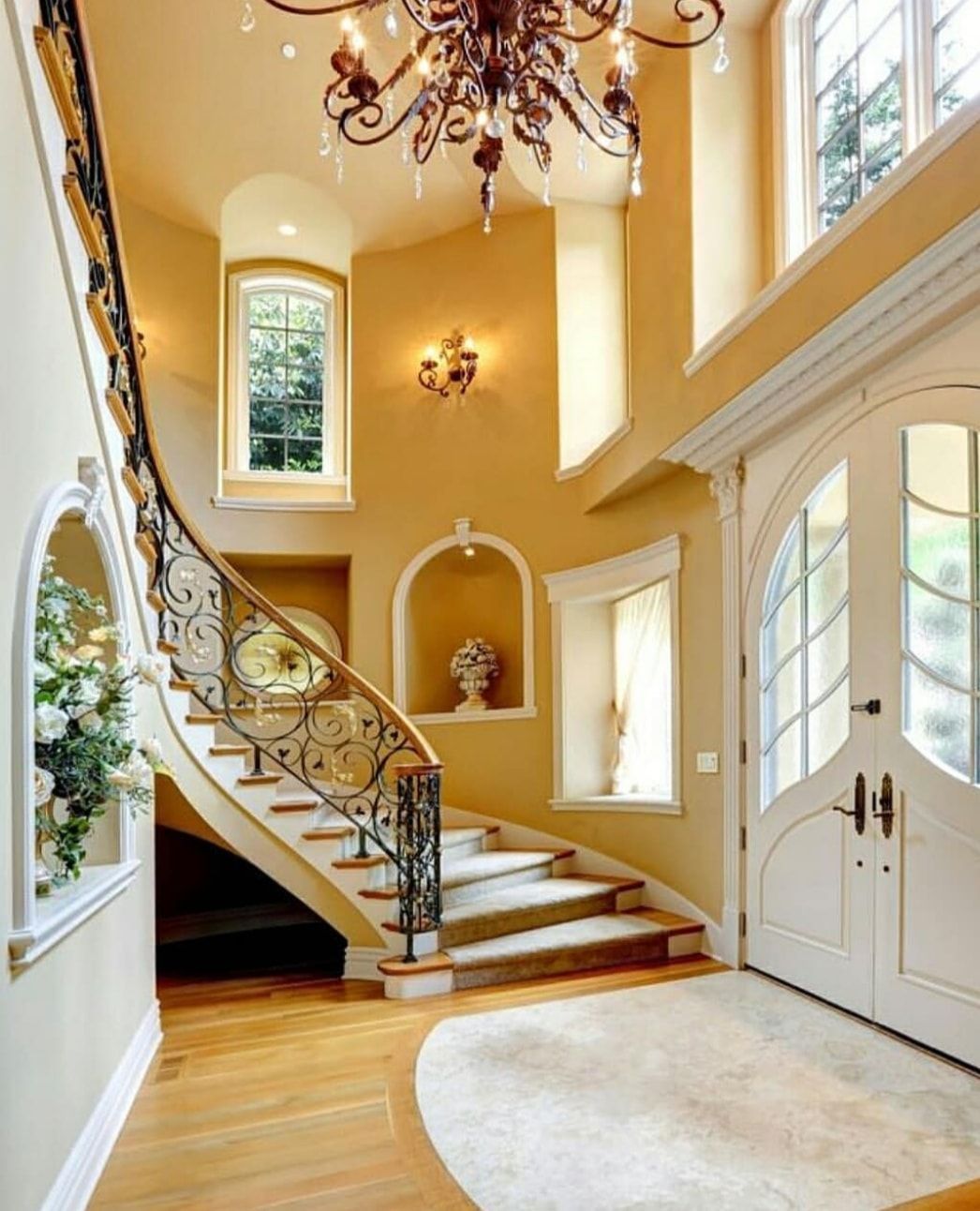 Elegant foyer with curved staircase, chandelier, tall windows, and double front doors on a polished wood floor.