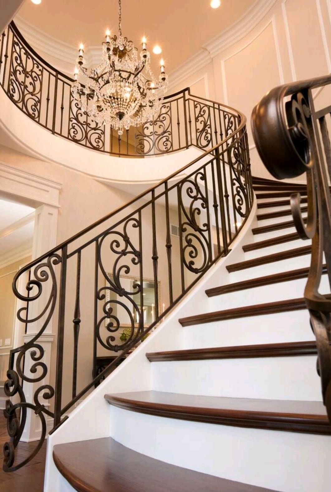 Elegant curved staircase with ornate black iron railings and a chandelier in a bright foyer