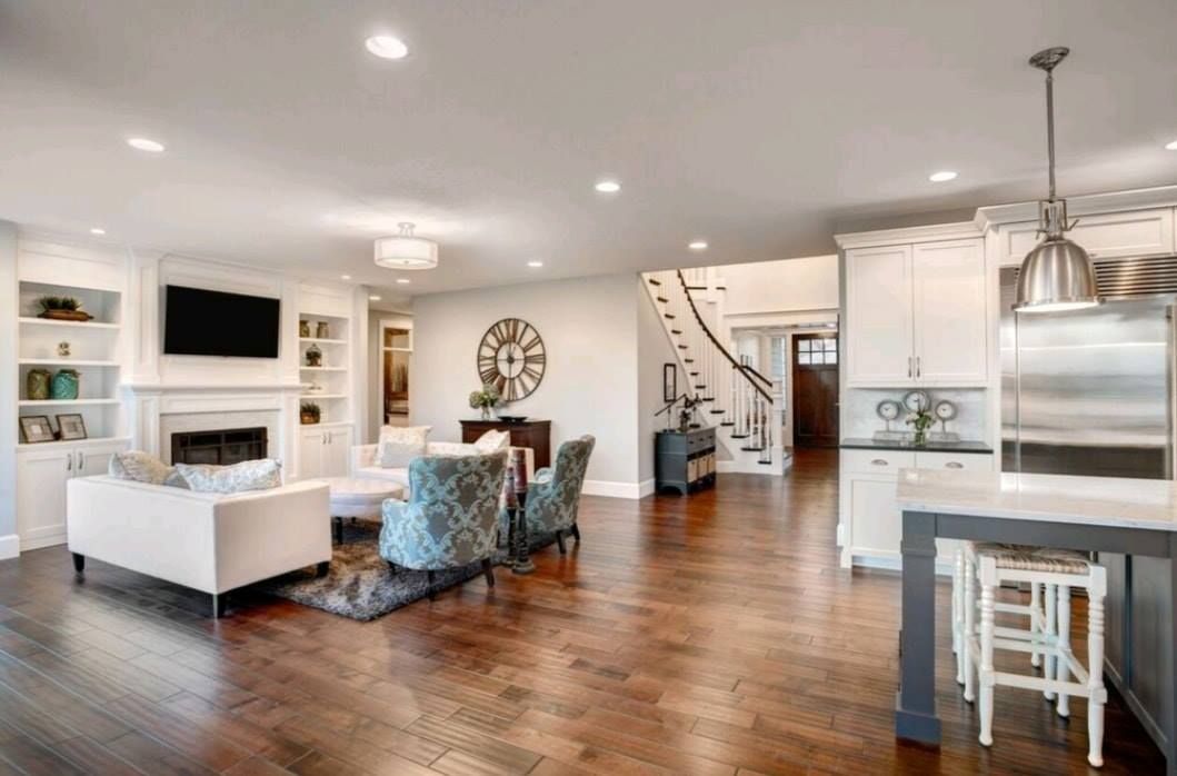 Bright open-concept living room and kitchen with white furniture, wood floors, and staircase in the background