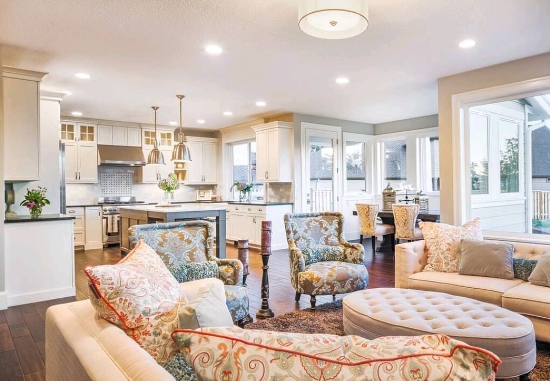 Bright open living room with floral sofas, tufted ottoman, and a kitchen in the background