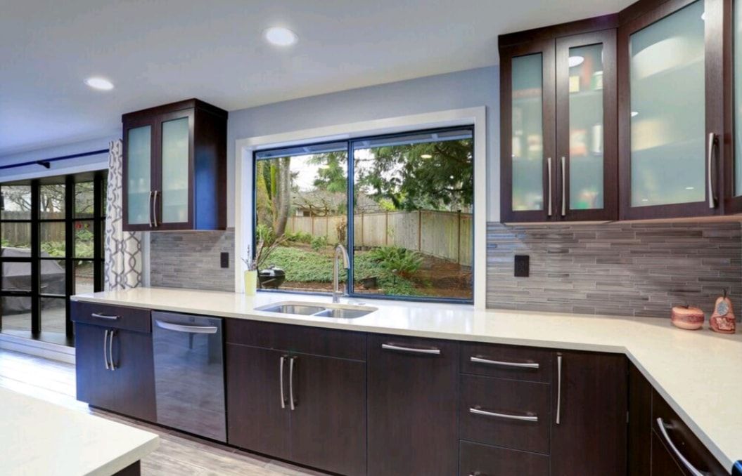 Modern kitchen with dark cabinets, white counters, and a large window overlooking greenery.