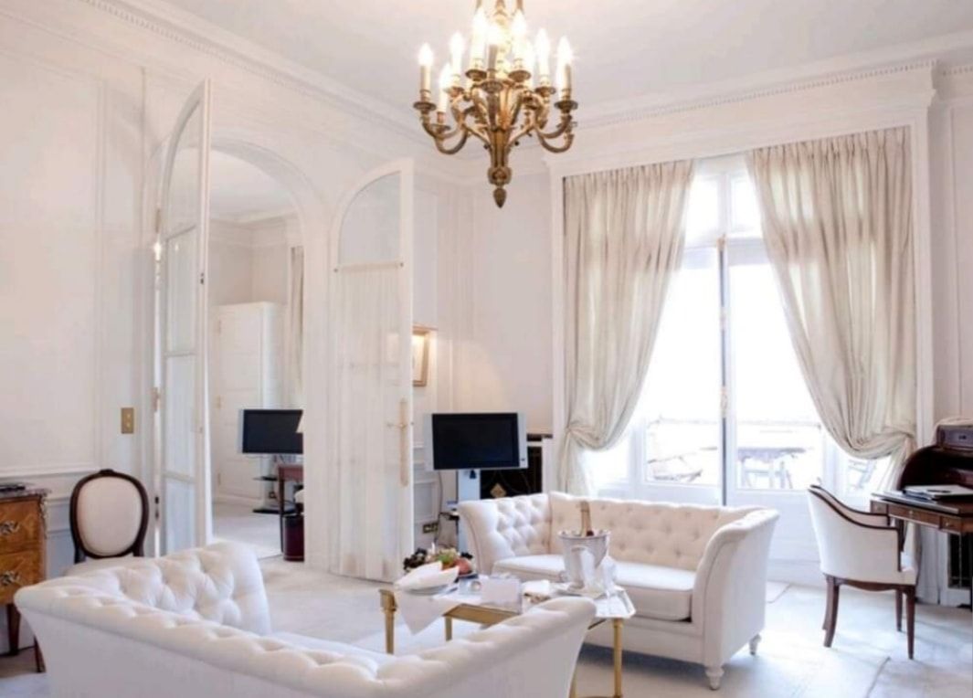 Elegant white living room with chandelier, tufted sofas, large windows, and soft curtains