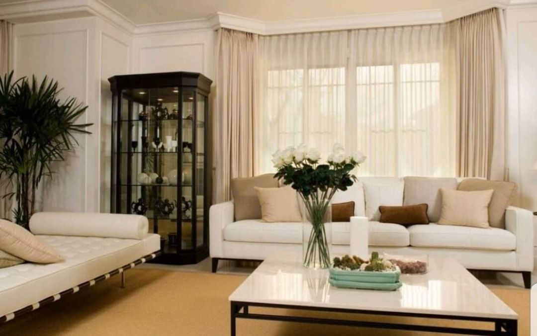 Bright living room with cream sofas, glass coffee table, display cabinet, and tall windows with curtains