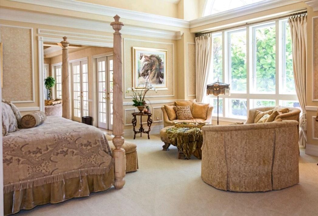 Elegant cream bedroom with a canopy bed, upholstered chairs, and large windows letting in bright daylight