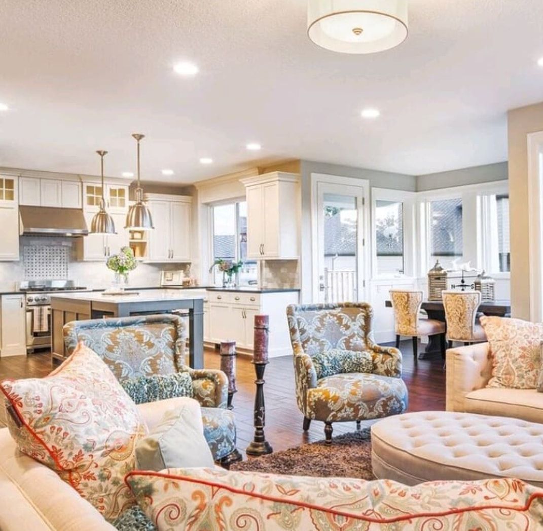 Bright open-concept living room and kitchen with patterned chairs, tufted ottoman, and pendant lights.