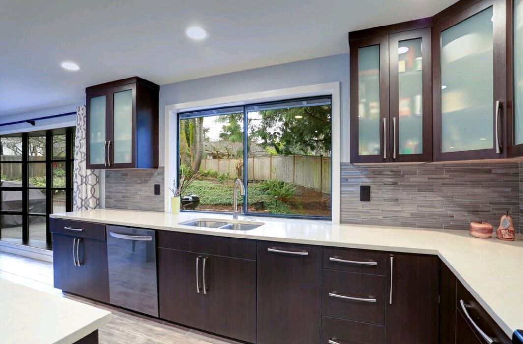 Modern kitchen with dark cabinets, light countertops, a large window, and glass-front upper cabinets.