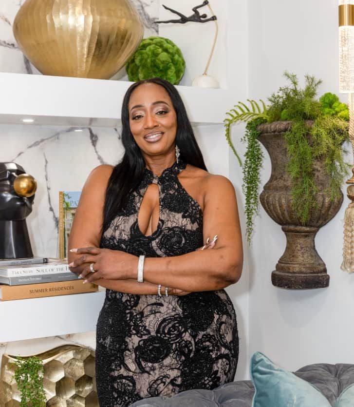 Woman in a black lace dress posing indoors beside a decorative vase and plant arrangement
