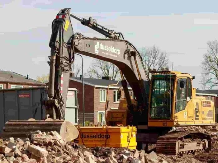 Excavator on a construction site beside piles of rubble and a brick building under demolition