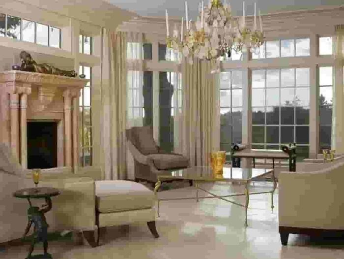 Bright living room with fireplace, chandelier, large windows, and beige chairs around a glass table