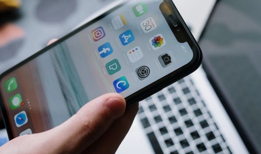 A close-up of a hand holding a smartphone displaying a home screen with various app icons, with a laptop in the background.