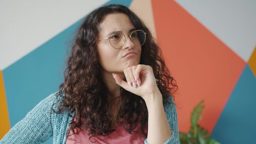 A person with curly hair and glasses rests their chin on their hand, looking upward with a thoughtful, skeptical expression.