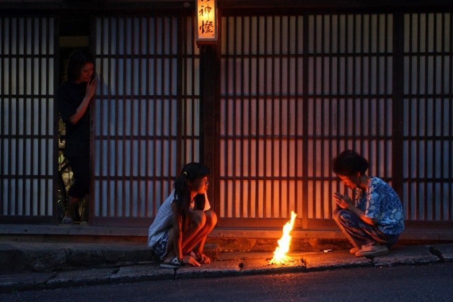Three people sit by a small fire on a dim street at night, in front of a closed shuttered storefront.