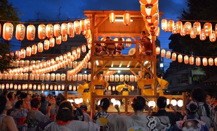 Crowded outdoor festival with glowing lanterns and a brightly lit wooden stage at dusk