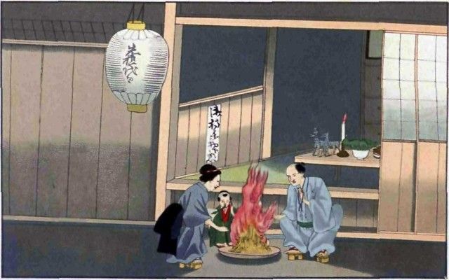 Two people sit by a small fire outside a wooden building at night, lit by a hanging lantern.