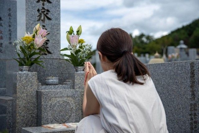 Person kneels at a cemetery grave, holding prayer hands beside white flowers and a stone marker.