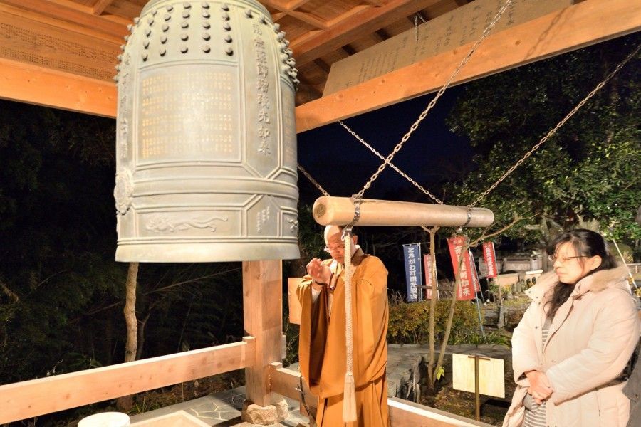 A monk strikes a large temple bell at night, while a person watches nearby.