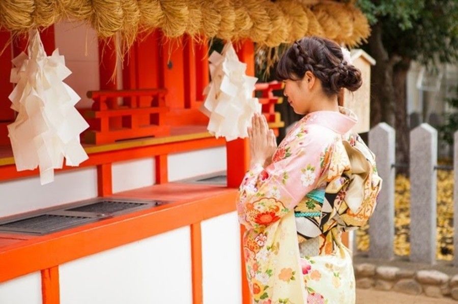 Woman in kimono praying at a shrine, hands clasped. Red and white structure with paper offerings.
