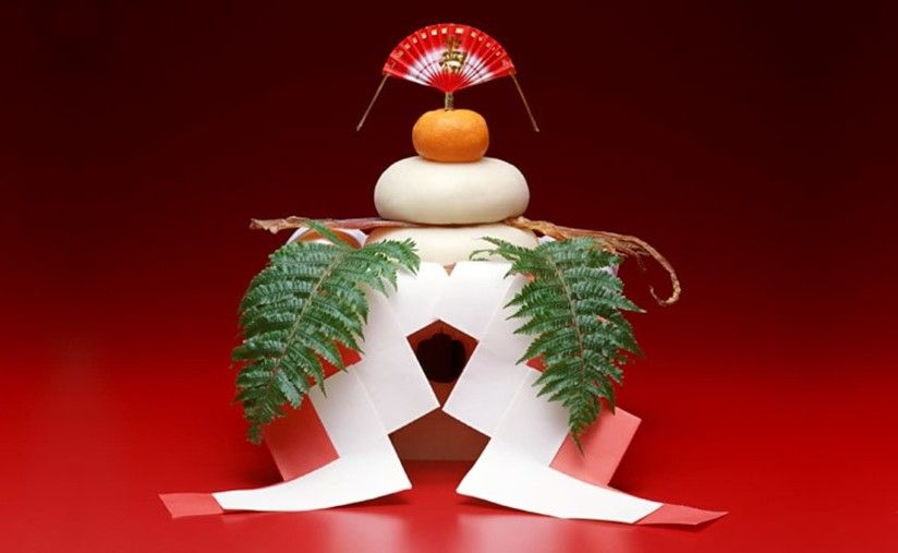 New Year’s Day mochi decoration: layered rice cakes, orange, fan, and paper embellishments on red background.