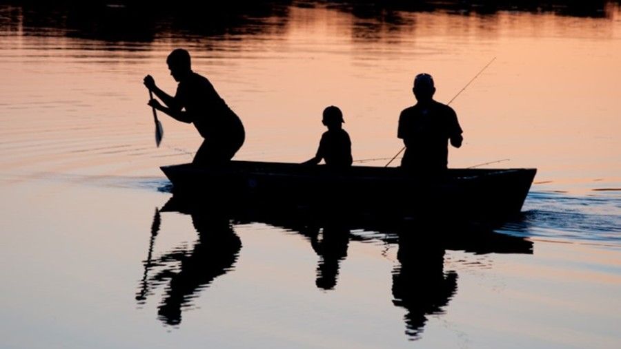 Three people are fishing in a boat on a lake at sunset.