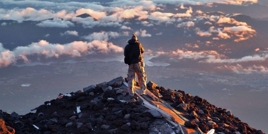 A man is standing on top of a mountain looking at the clouds.