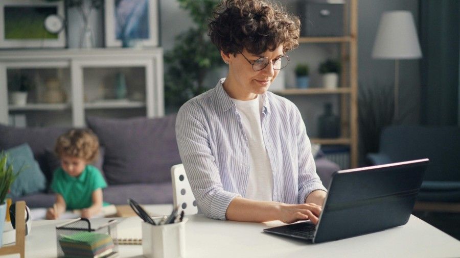 Woman with glasses working on laptop at table; child drawing in background.