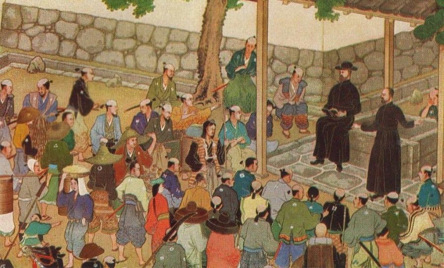 Japanese people gathered to hear two men in black robes speak; outdoor setting.