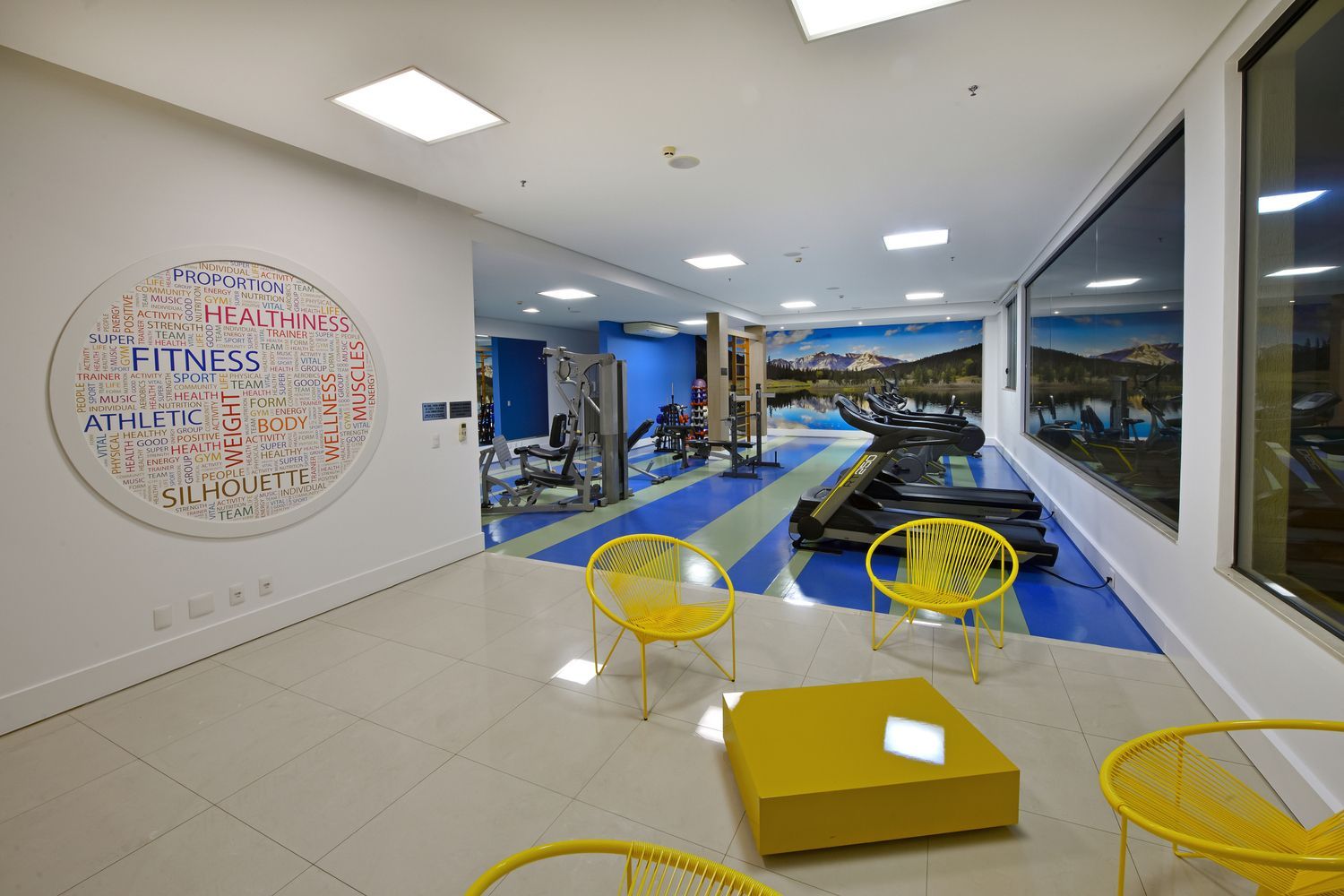 Uma academia com cadeiras amarelas, uma mesa e uma esteira.