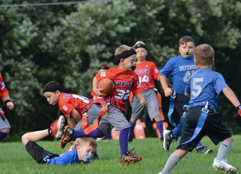 Flag football game at night, a player in red jersey runs with the ball as others pursue on a green field.