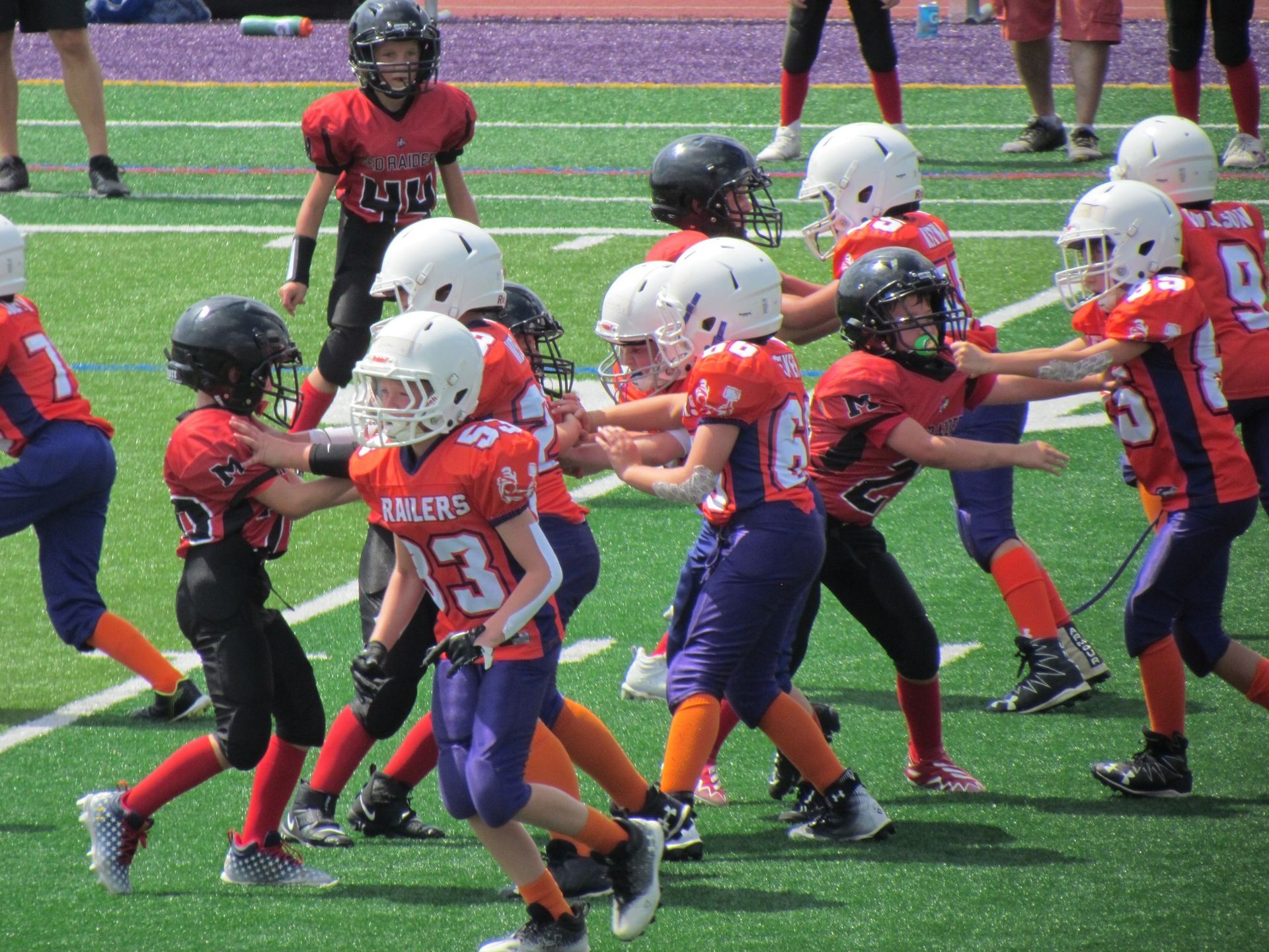 Football players in action on a grassy field; one runs with the ball, pursued by two opponents.