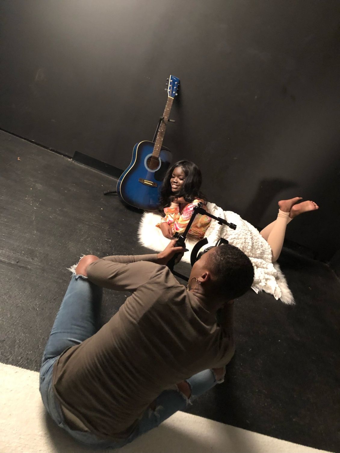 A man is taking a picture of a woman laying on the floor with a guitar in the background