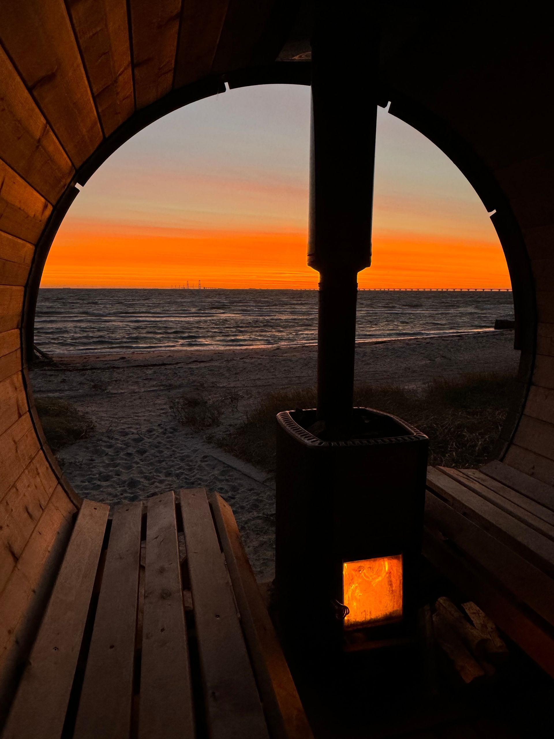 Sonnenuntergangsblick durch ein Saunafenster: Meer, orangefarbener Himmel, brennender Ofen, hölzernes Interieur.